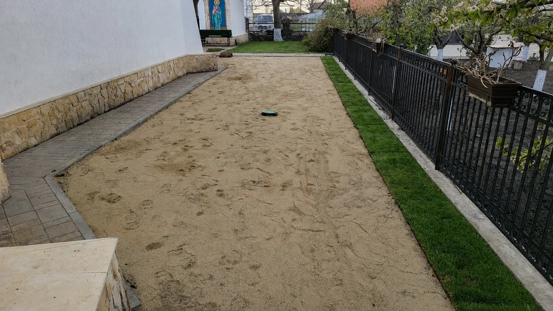 Pat de nisip nivelat, pregătit pentru gazon, între o alee pavată și o fâșie de iarbă deja prinsă lângă gard